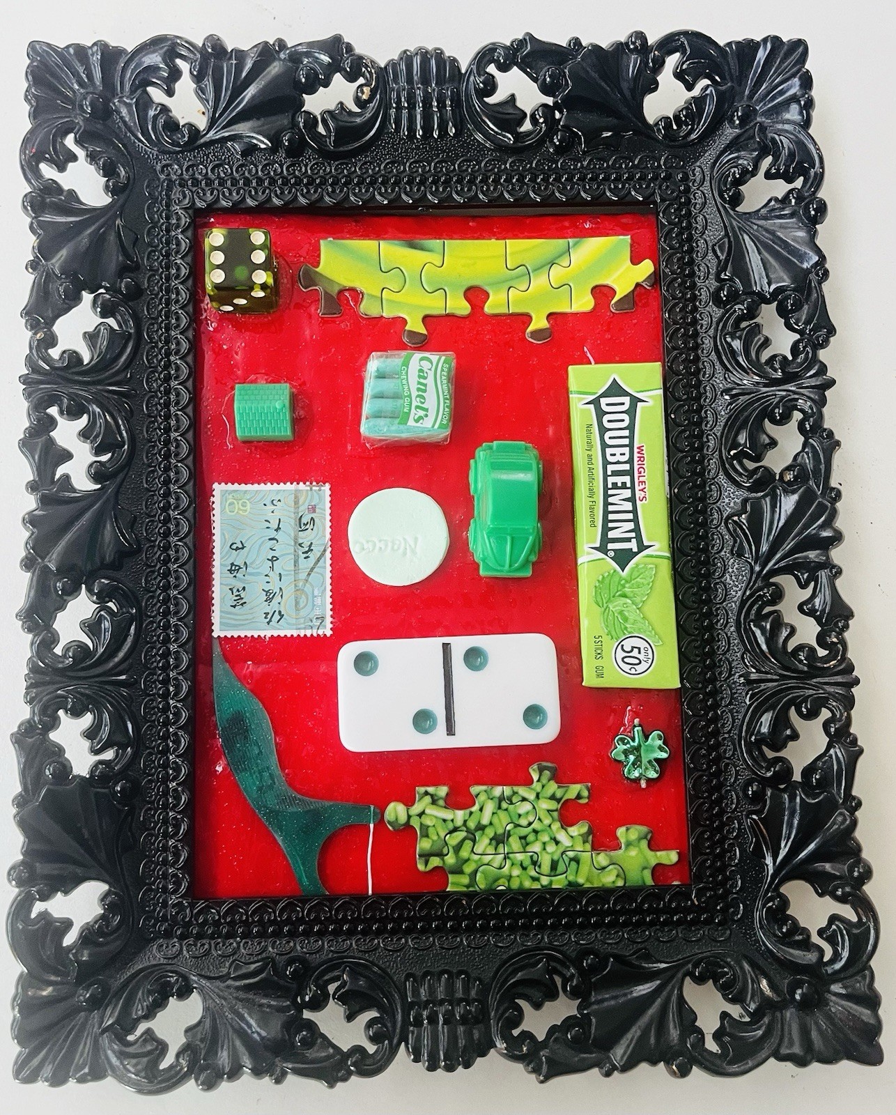 Original art: found green objects on red background in ornate brown frame. 7x9”
